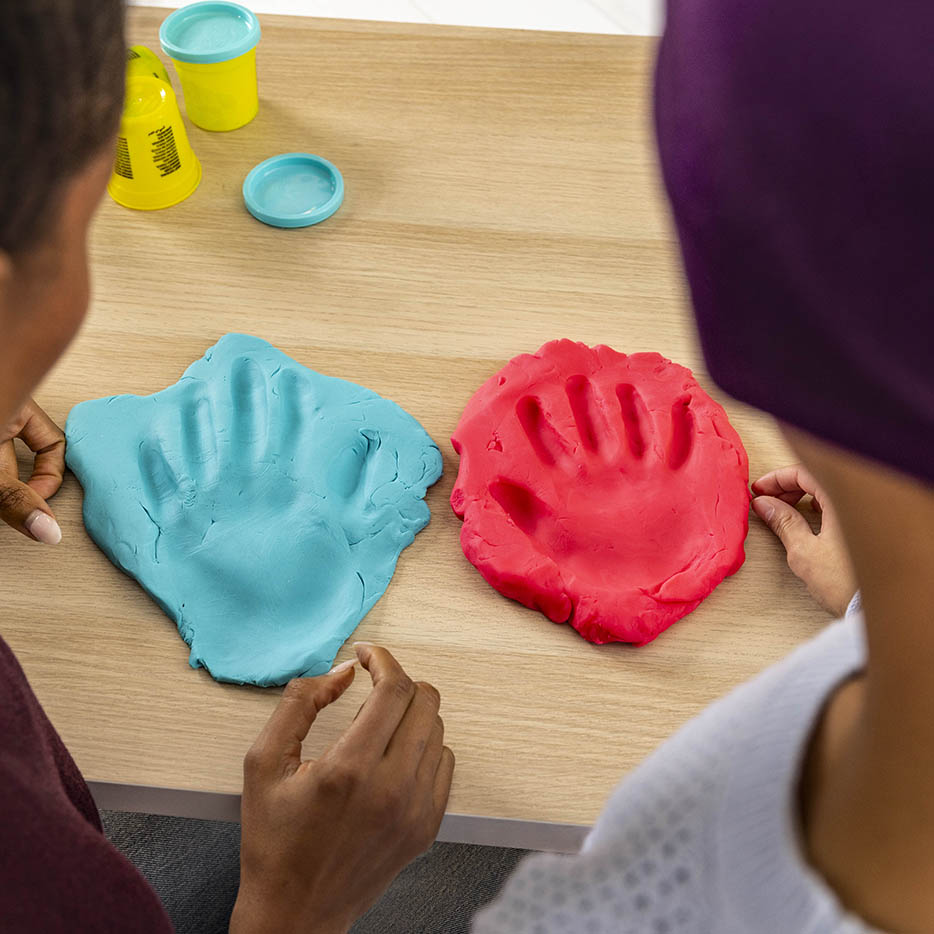 An overhead shot shows two handprints, one in blue clay and one in red clay, pressed onto a light wood table with two people's hands nearby.