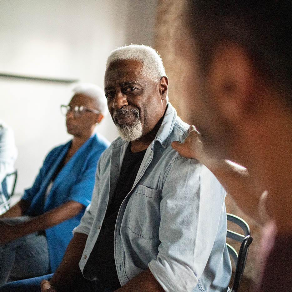 A friendly, silver-haired Black man looks confidently at the camera while receiving a reassuring touch on the shoulder.
