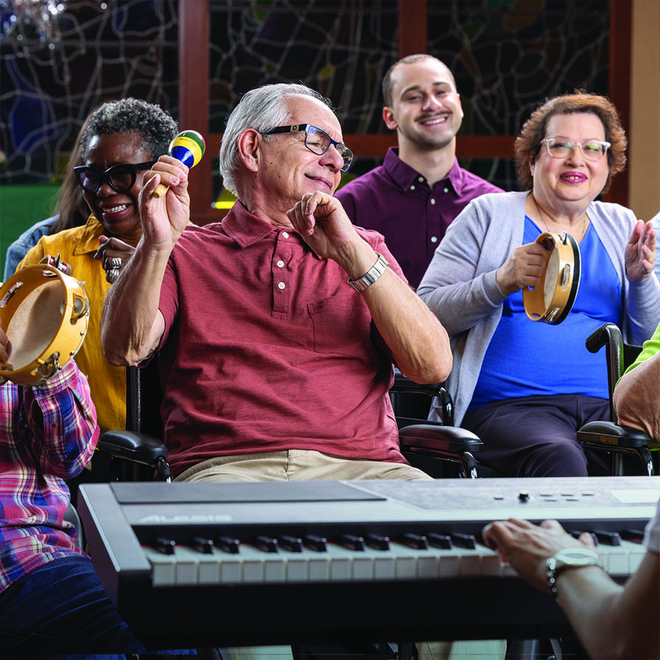 A diverse group of happy people, including seniors in wheelchairs, plays musical instruments like a maraca, tambourines, and a keyboard.