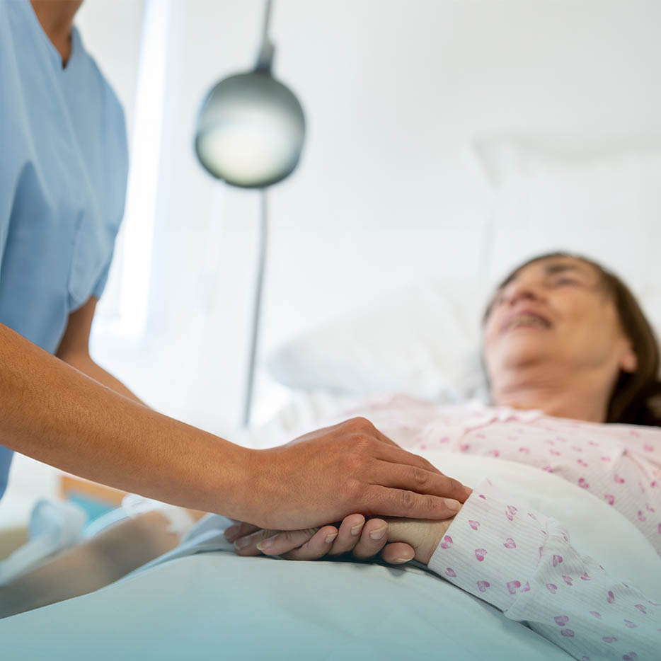 A nurse, wearing blue scrubs, gently holds the hand of an elderly, smiling woman who is lying in a hospital or home care bed.