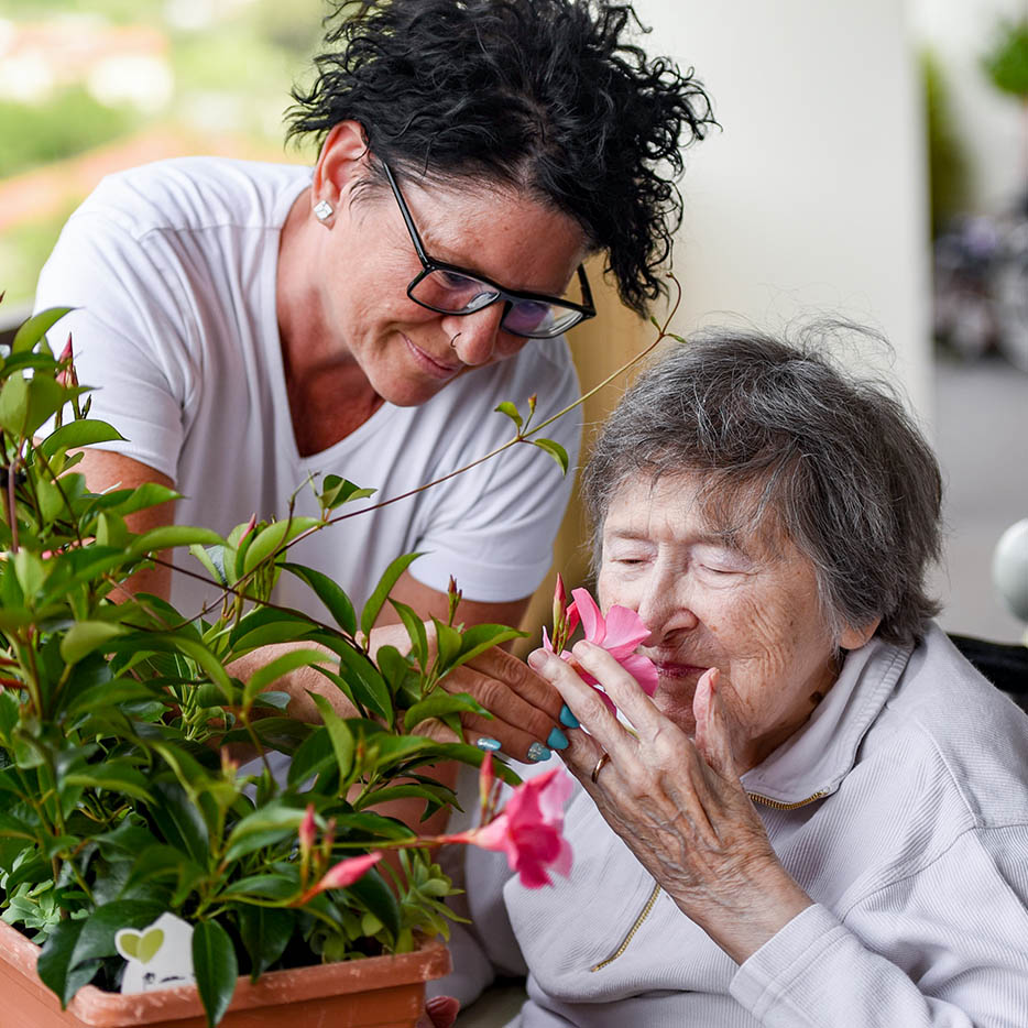 A caregiver helps an elderly woman with her eyes closed smell a pink flower from a potted plant on an outdoor patio.