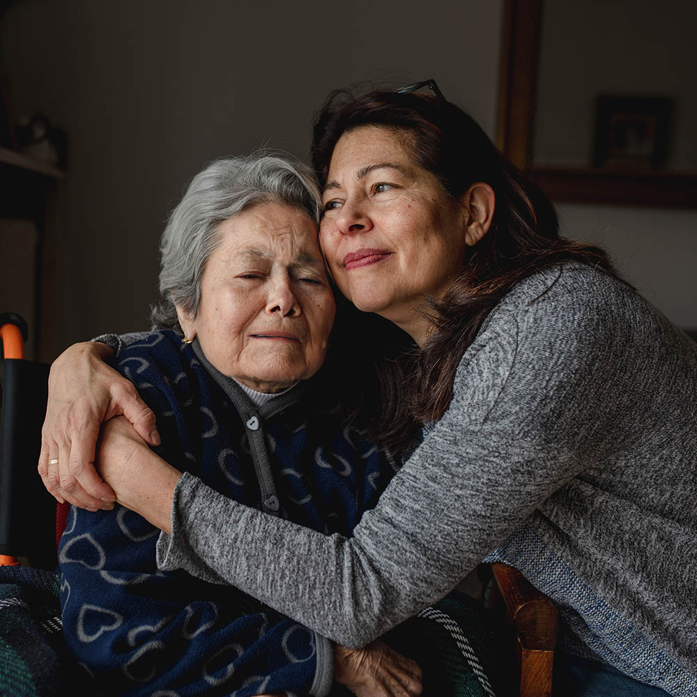 A woman with long dark hair leans her cheek against an elderly woman's head, embracing her warmly in a moment of shared comfort and affection.