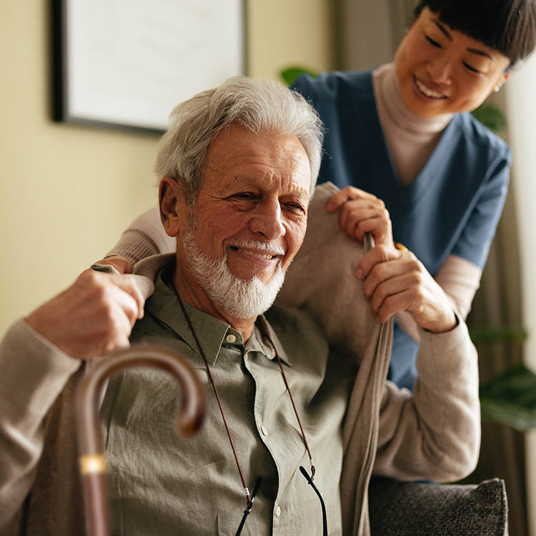 A smiling caregiver helps a senior man with a white beard put on a cardigan.