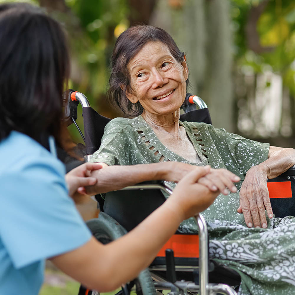 An elderly woman in a wheelchair smiles warmly while a caregiver in blue scrubs holds her hand outdoors.