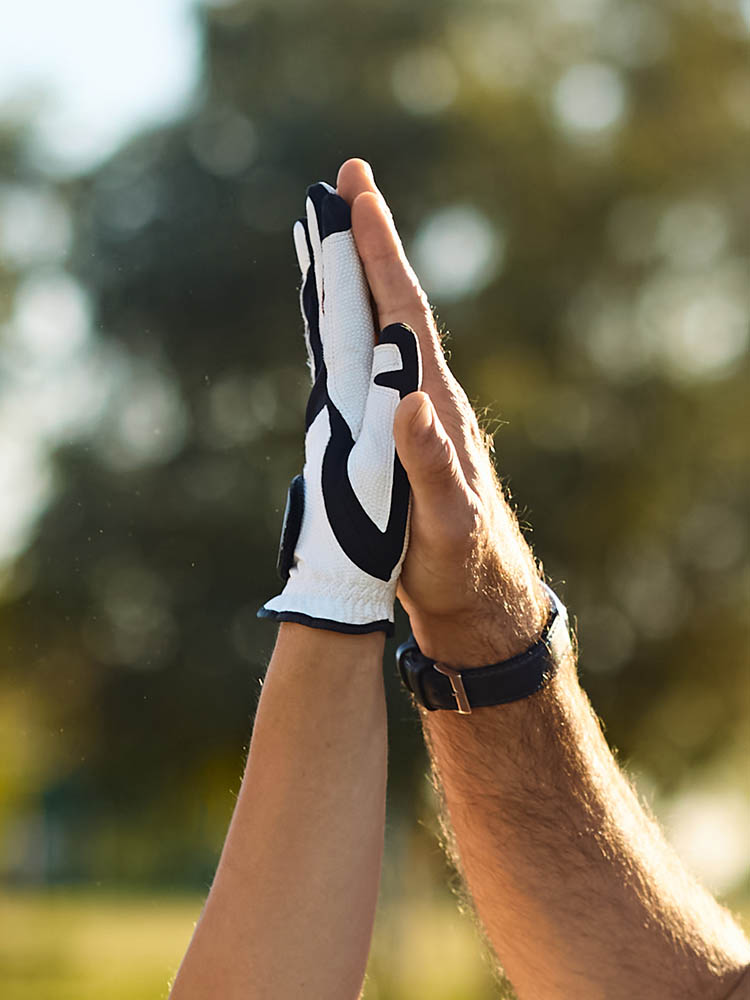 A close-up of two people giving a high five outdoors, with one person wearing a white and black golf glove against a blurred green background.