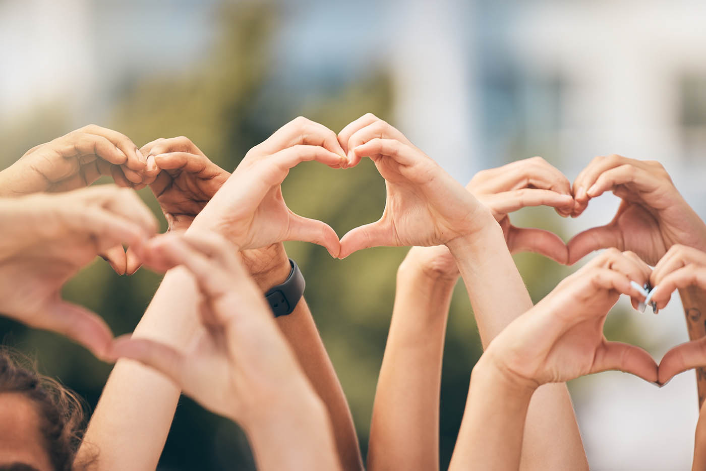 A close-up of multiple sets of hands raised against a blurred green background, with fingers joined together to form several heart shapes.