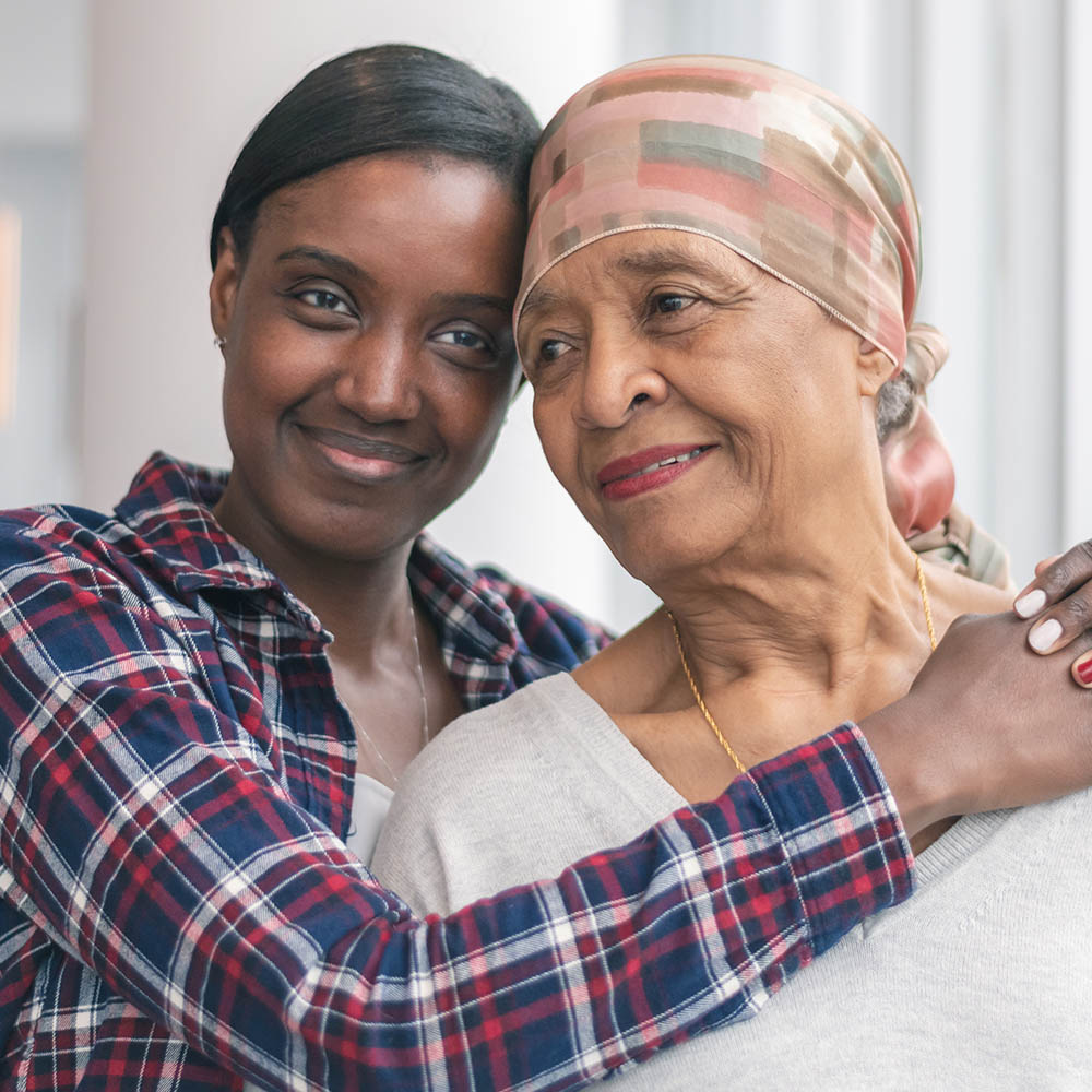 A young woman in a plaid shirt affectionately embraces an older woman wearing a headscarf, both smiling gently.