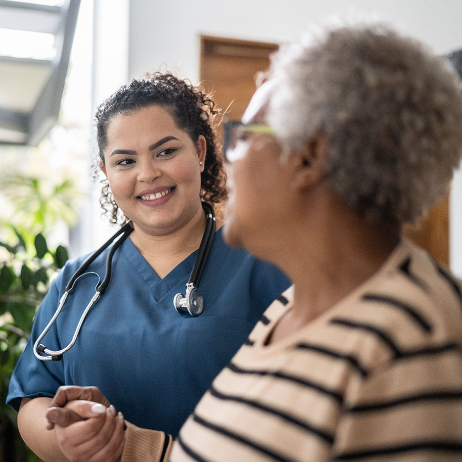 A smiling female nurse in blue scrubs and a stethoscope holds an elderly woman's hand in a bright room.