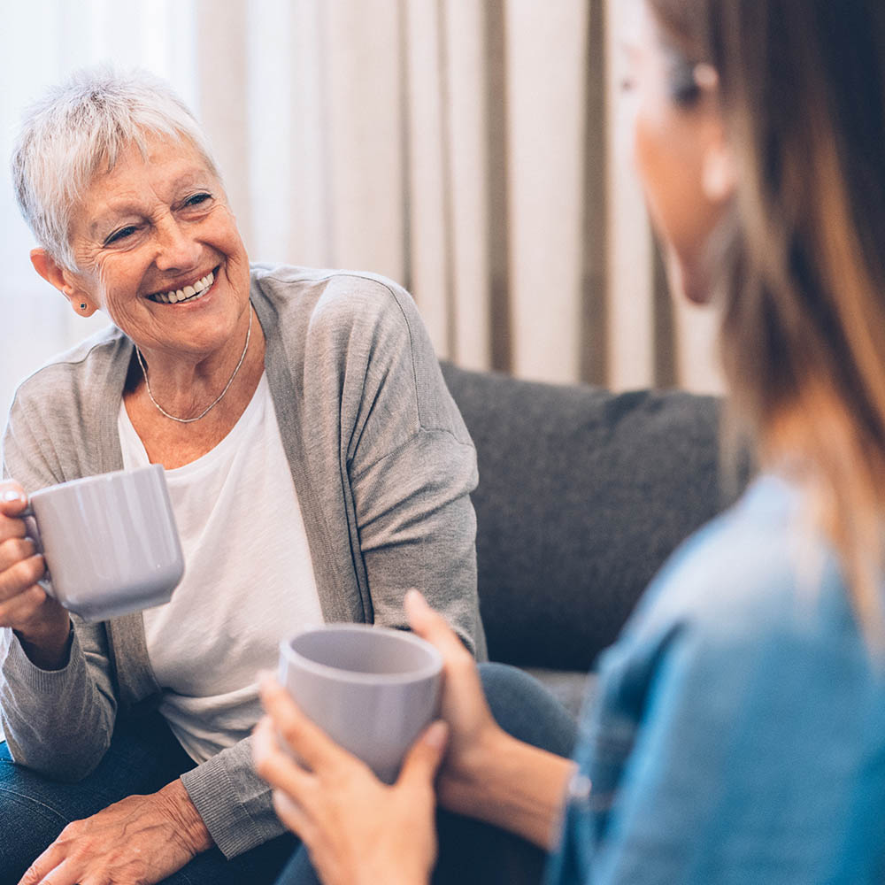 A senior woman with short gray hair smiles warmly while holding a mug and conversing with a younger woman over coffee.