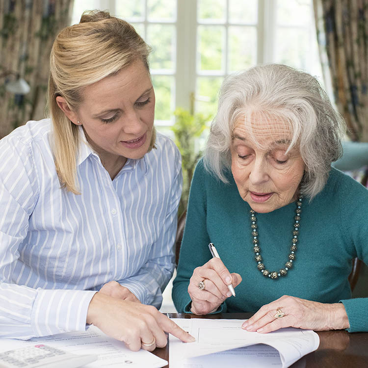 Older woman signs paperwork at a table while a younger woman beside her points and helps review the documents.