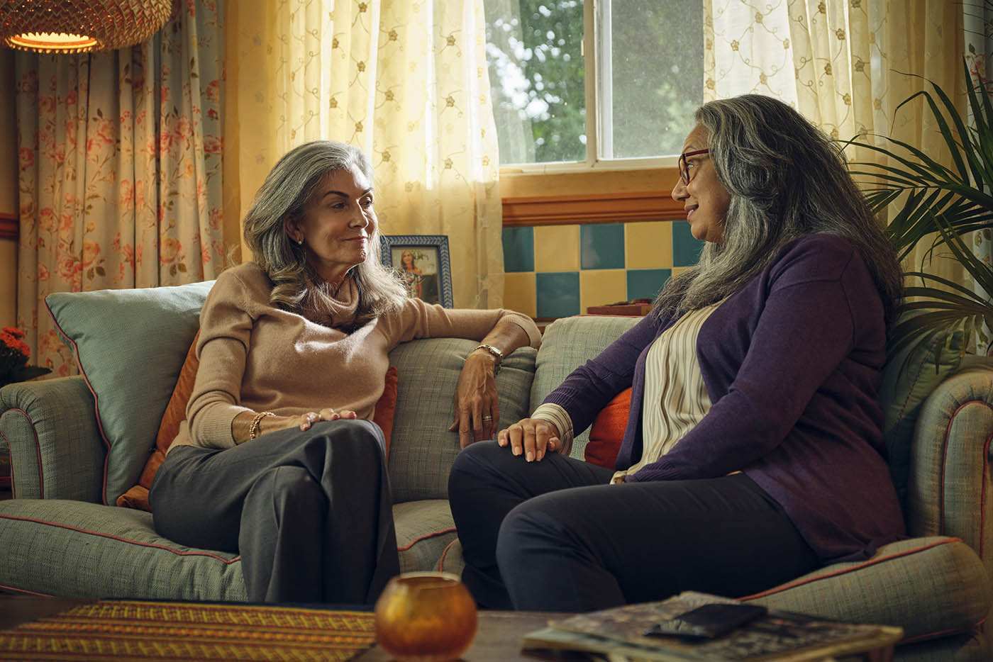 Two senior women with long gray hair sit together on a comfortable sofa in a brightly lit living room, engaged in a peaceful conversation.
