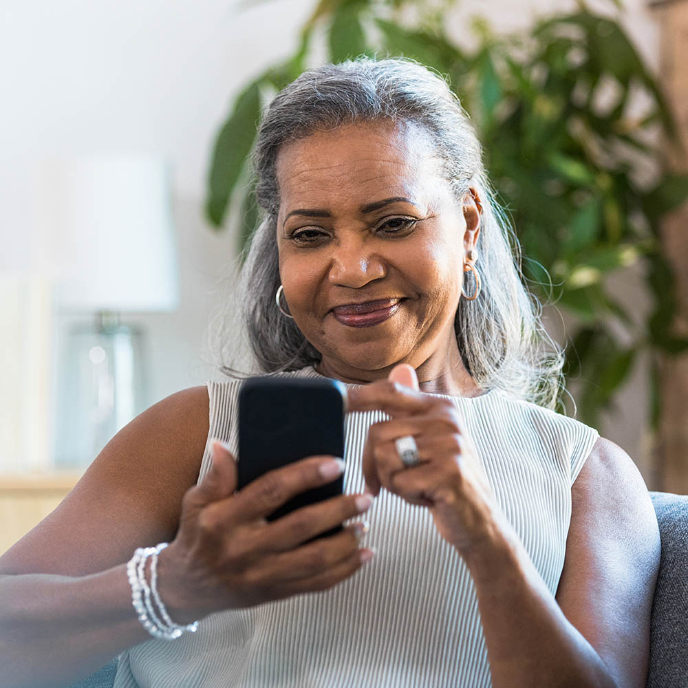 A senior woman with grey hair and hoop earrings smiles while looking down at her smartphone, which she is holding and using with one hand.