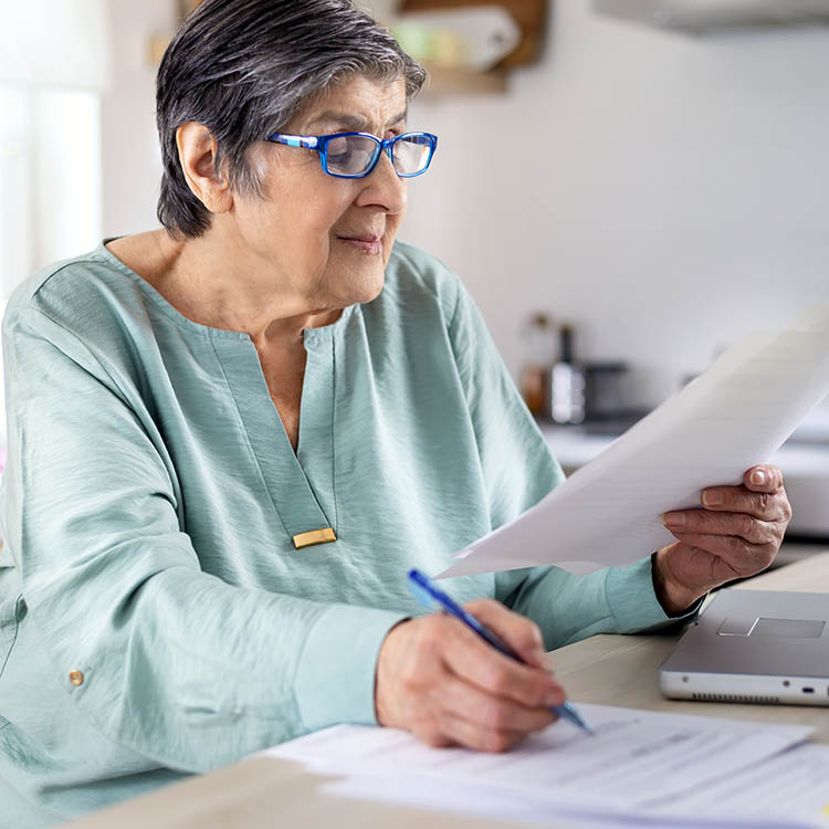 Senior woman in blue glasses writing on documents at a desk with a laptop.